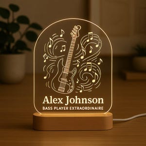 Może przedstawiać: Podświetlana lampka nocna w kształcie gitary basowej z imieniem "Alex Johnson" i tekstem "Bass Player Extraordinaire" wytrawionym w akrylu. Światło stoi na drewnianej podstawie i zawiera nuty.