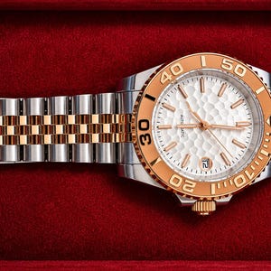 May include: A luxury wristwatch with a silver and gold-tone stainless steel strap and a white face. The watch has a gold-tone bezel with black markings and a date display. The watch is presented on a red velvet surface.