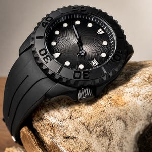 Könnte beinhalten: Eine mattschwarze Armbanduhr mit einem strukturierten Zifferblatt mit Wirbelmuster, leuchtenden runden Stundenmarkierungen und einem Datumsfenster auf der Drei-Uhr-Position. Die Uhr hat eine drehbare Lünette mit eingravierten Ziffern und ein schwarzes Kautschukarmband.