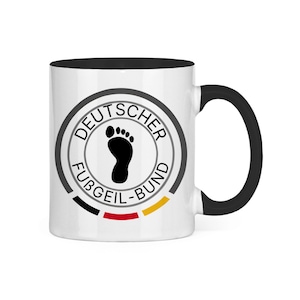 Op de afbeelding: Een witte keramische mok met een zwarte handgreep en binnenkant. De voorzijde toont een cirkelvormig logo met de tekst DEUTSCHER FUBGEIL-BUND, een zwarte voetafdruk in het midden en een horizontale zwarte, rode en gele streep onderaan.