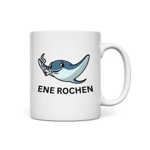 May include: White ceramic mug with a cartoon stingray smoking a cigarette. The stingray is light blue and white with a smiling face. The text "ENE ROCHEN" is printed in black below the stingray.