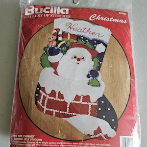 May include: A Bucilla Gallery of Stitches Christmas felt stocking kit titled Down the Chimney. The packaging features a graphic of Santa Claus emerging from a brick chimney, a teddy bear, and the name Heather. It is a 15 inch diagonal kit.