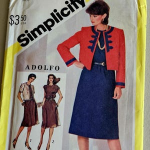 May include: Simplicity pattern envelope featuring a woman in a red jacket and navy blue dress. The envelope includes the text "Simplicity" and "ADOLFO", along with the pattern number 5486 and size 12.