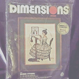 May include: A Dimensions crewel embroidery kit titled Woman Stitching, featuring a Norman Rockwell illustration of a person in a ruffled dress working at a frame. The plastic packaging displays the 12 by 16 inch frame size and kit contents.