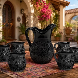 May include: A black ceramic pitcher and five matching mugs featuring a raised, glossy scrollwork pattern. The set is arranged on a patterned textile. Each piece has a rounded body, a sturdy handle, and a smooth, dark finish with intricate details.