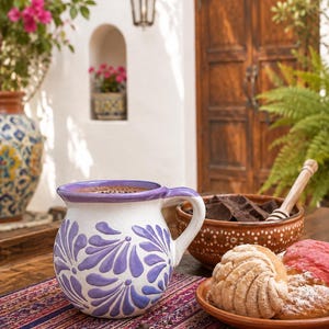 May include: A round ceramic mug with a white base and purple painted floral patterns, featuring a purple rim and handle. Beside it sits a brown clay bowl filled with dark chocolate chunks and a plate containing two sweet bread rolls with textured tops.