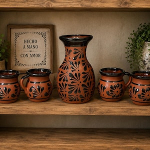 Puede incluir: Una jarra de terracota y cuatro tazas a juego con un patrón floral negro pintado a mano. La jarra tiene un borde ancho y cuerpo redondeado. Cada taza incluye un asa. Un letrero enmarcado muestra el texto HECHO A MANO CON AMOR.