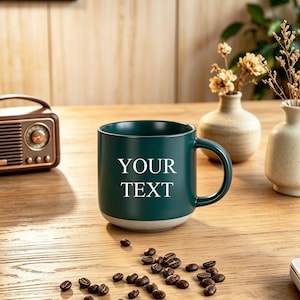 May include: A dark green ceramic mug with a light gray base featuring the white text "YOUR TEXT" printed on the front. The mug has a smooth finish and a curved handle, positioned on a wooden surface with scattered dark roasted coffee beans.