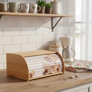 Puede incluir: Una panera de bambú de color marrón claro con tapa enrollable. La parte delantera de la panera presenta una imagen impresa de pan. La panera está sobre una encimera de madera en una cocina.