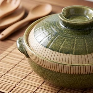 Puede incluir: Una olla donabe japonesa de cerámica verde con tapa. La olla tiene una superficie texturizada y una banda beige claro con líneas verticales. Cucharas de madera en el fondo. La olla está sobre una esterilla de bambú.