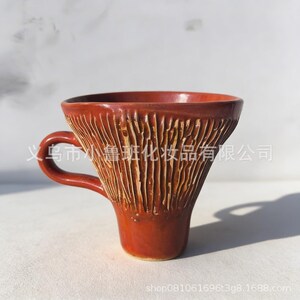 Puede incluir: Una taza de cerámica de color marrón rojizo con un exterior texturizado y tallado. La taza tiene un asa y una parte superior ancha y acampanada. La base es de un tono marrón más oscuro. La taza está sobre una superficie blanca.