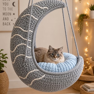 May include: A gray, crescent-shaped cat bed with white accents and a light blue cushion. A fluffy cat with blue eyes rests inside the bed, which hangs from a rope. The text on the wall says "Love you to the moon and back."