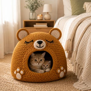 Puede incluir: Una cama para gatos hecha a mano, de color mostaza, con forma de cabeza de oso. La cama presenta una cara de oso blanca y negra, ojos cerrados y huellas de patas. Dos gatos están acurrucados dentro de la acogedora abertura redonda.