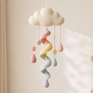 Op de afbeelding: Een gehaakte hangende mobiel met een grote crèmekleurige wolk aan de bovenkant. Aan de wolk hangen vier draden met kleine sterretjes en druppelvormen in pastelroze, geel en blauw, plus een centrale spiraal in een regenboogverloop.