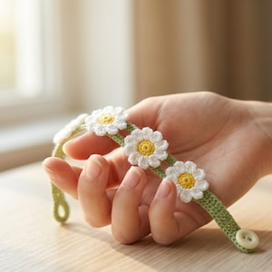 May include: A handmade crochet daisy chain bracelet. The bracelet features white daisies with yellow centers, attached to a green band. A white button secures the bracelet. The bracelet is held in a hand.