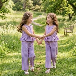 Puede incluir: Dos niñas pequeñas con conjuntos a juego de cuadros morados y blancos, tomadas de la mano y sonriendo. Los conjuntos incluyen tops sin mangas con detalles de peplum y pantalones de pernera ancha. Llevan sandalias blancas.