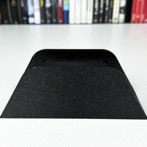 May include: A black, textured, angled stand with a rectangular top. The stand has a grainy appearance and is designed to hold a small electronic device. The stand is made of a solid material and sits on a white surface.