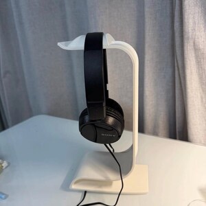 Può includere: Cuffie Sony nere over-ear appese a un supporto bianco per cuffie. Il supporto ha un design curvo con una base e un alloggiamento superiore per le cuffie. Le cuffie sono collegate a un cavo nero.
