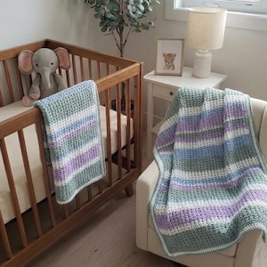 Peut inclure: Un berceau en bois avec un éléphant en peluche gris et une couverture en crochet à rayures dans les tons de vert, violet, bleu et blanc. Une autre couverture est drapée sur une chaise. Une lampe et une image encadrée sont sur une table de chevet.