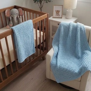 Peut inclure: Une couverture de bébé bleu clair, crochetée, drapée sur un berceau en bois et un fauteuil blanc. Un éléphant en peluche gris est assis dans le berceau. La couverture a un motif tissé texturé. Une image encadrée et une lampe sont sur une table de chevet.