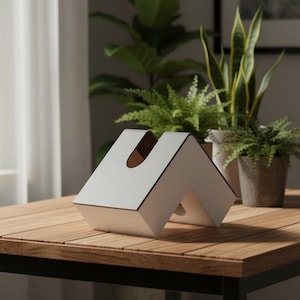 Puede incluir: Un objeto blanco, angular y en forma de M con un acabado liso y bordes oscuros. La estructura presenta un recorte central y un interior hueco, formando una silueta geométrica similar a una tienda de campaña sobre una superficie plana.