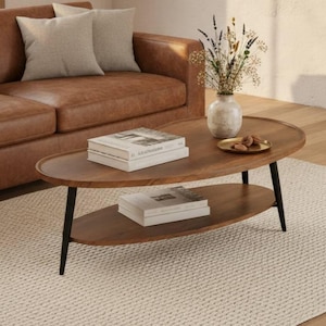 May include: An oval coffee table with a dark wood top and a matching lower shelf, supported by four thin black metal legs. The table holds two stacked books on the top surface, a small ceramic vase with dried flowers, and a gold tray with items.