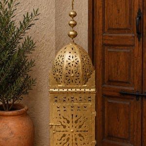 Può includere: Una lanterna in metallo color oro con intricati disegni traforati. La lanterna ha una parte superiore a cupola e una base rettangolare con una piccola porta. Il design comprende motivi floreali e geometrici. La lanterna è accanto a una porta di legno marrone.