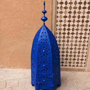May include: A tall, cobalt blue metal lantern featuring an intricate, repeating star-shaped cutout pattern across its arched body. The lantern is topped with a decorative finial and a small hanging loop, and it includes a hinged door at the base.