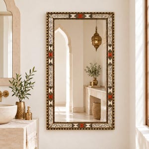 Può includere: Uno specchio da parete rettangolare con una cornice decorata caratterizzata da intricati motivi a volute nere, accenti bianchi e inserti geometrici rossi a forma di diamante al centro di ogni lato e negli angoli della cornice.