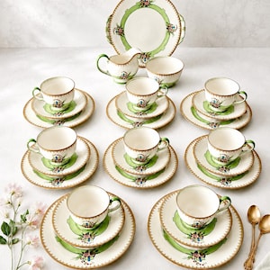 Può includere: Un servizio da tè in ceramica con base color crema, bordo verde chiaro, finiture dorate e piccoli motivi floreali. Il set include un grande piatto da portata, una lattiera, una zuccheriera e otto coperti, ciascuno con tazza e due piattini.
