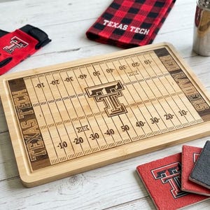May include: A rectangular bamboo cutting board laser-engraved with a football field design, featuring the Texas Tech logo, Red Raiders text, and yard line markings. The board is surrounded by a keychain, red and black gloves, a plaid towel, and coasters.