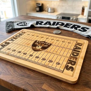 May include: A rectangular bamboo chopping board engraved with a football field design, the text LAS VEGAS RAIDERS, and the Raiders team logo. A grey and black knit scarf with the text RAIDERS and a team logo is positioned behind the board.