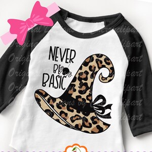 May include: A white and black raglan-style shirt with the words "NEVER BE BASIC" and a leopard print witch hat design. A black spider hangs from the text. The shirt has black sleeves and a white body.