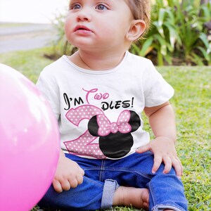 May include: A young child wearing a white t-shirt with pink polka dots and the text "I'm Two Dles!"  The child is wearing blue jeans and a pink bow in their hair. The child is sitting on a green grassy surface.