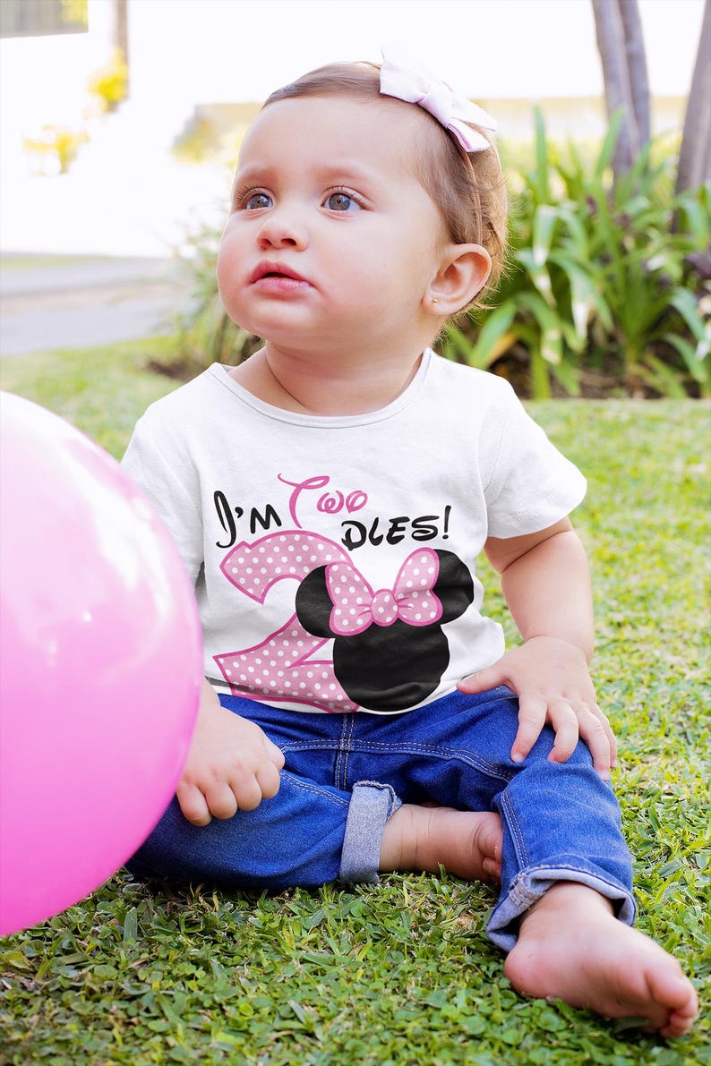 May include: A young child wearing a white t-shirt with pink polka dots and the text "I'm Two Dles!"  The child is wearing blue jeans and a pink bow in their hair. The child is sitting on a green grassy surface.