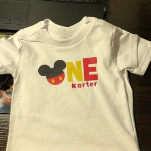 May include: White t-shirt with a Mickey Mouse head and the word "ONE" in yellow and red letters. The name "Karter" is printed below the word "ONE".