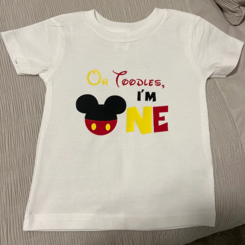 以下が含まれることがあります： ミッキーマウスの頭が描かれた白いTシャツ。赤と黒で、黄色と赤で「Oh Toodles, I'm One」の文字が書かれています。
