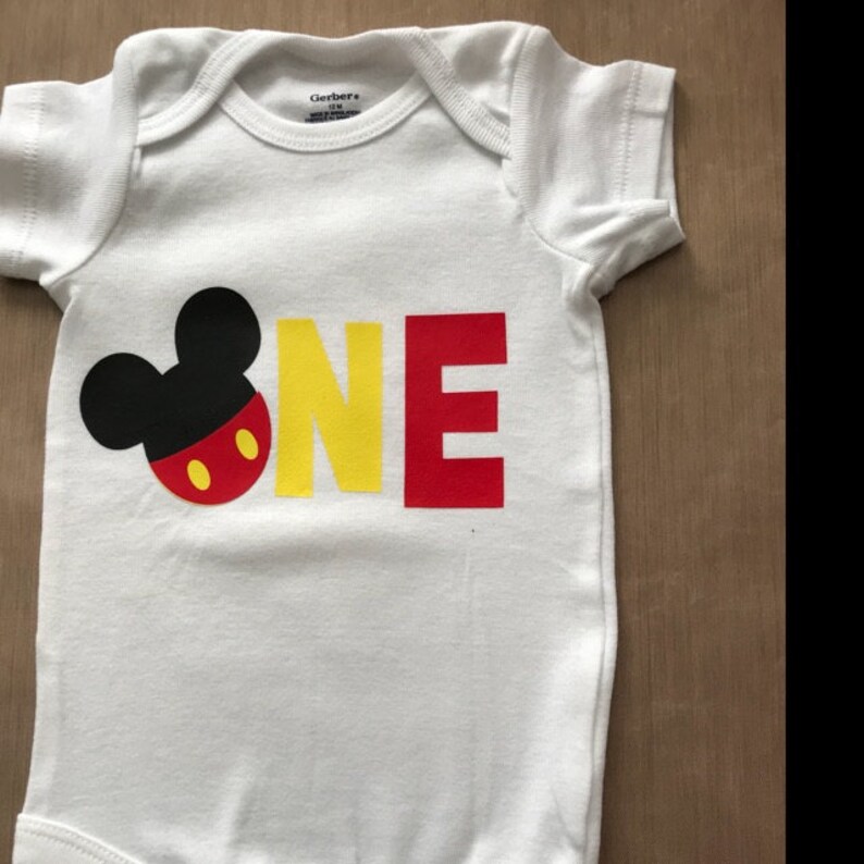 May include: White baby onesie with a black Mickey Mouse head and the word "ONE" in yellow and red letters.