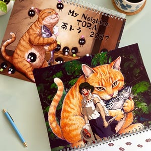 May include: A 2027 spiral-bound wall calendar titled My Neighbor TORA featuring illustrations of a large orange tabby cat holding a fish. One page shows the cat with small black soot sprites, and another shows a character standing before the cat.