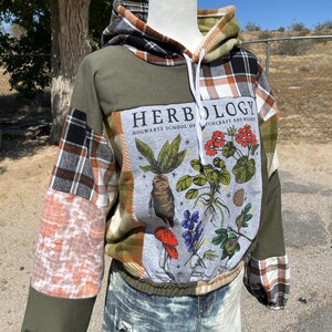 May include: Olive green and plaid patchwork hoodie with a botanical print. The print features illustrations of plants and the text "Herbology Hogwarts School of Witchcraft and Wizardry". The hoodie has a drawstring hood and elastic waist.