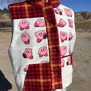 May include: A sleeveless vest featuring a red and yellow plaid pattern on the collar, front placket, and lower pocket, contrasted with white panels printed with multiple small pink Kirby character graphics and secured with metallic snap buttons.