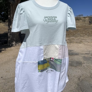 May include: A light blue t-shirt with puff sleeves and a round neckline. The chest features the text "JACKSON HOLE WYOMING". The lower section is a white patchwork design with a graphic print of a yellow vehicle and the text "FRAME'S FINEST".