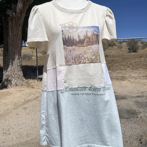 May include: A cream-colored short-sleeve shirt with puff sleeves and a color-blocked design. The front features a rectangular graphic of a mountain landscape with wildflowers and the text Mountain town classic WHERE THE WEST STAYS WILD.