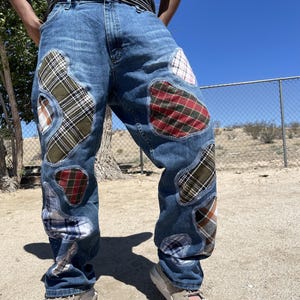 May include: Blue denim jeans featuring multiple irregular patches of various plaid and checkered fabrics in shades of red, green, white, and brown. The patches are stitched onto the legs with visible white thread, creating a textured appearance.