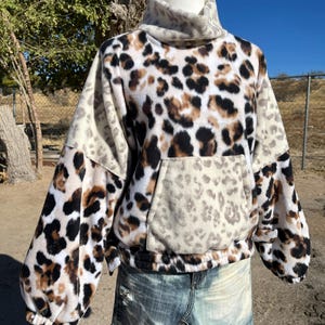 May include: A cozy fleece pullover with a leopard print design. The top features a high neck, long sleeves, and a kangaroo pocket. The color palette includes shades of brown, black, and cream.