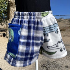 May include: Cozy fleece shorts with a blue plaid pattern on one side and a white background with alligator illustrations on the other. Features a blue pocket with a frog design. Elastic waistband.