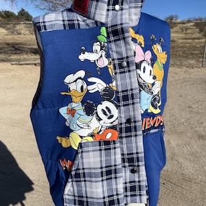 May include: A blue vest featuring a graphic print of Disney characters including Mickey Mouse, Donald Duck, Goofy, Minnie Mouse, and Pluto. The vest has a grey and navy blue plaid pattern on the collar, front placket, and pockets, with metal snap buttons.