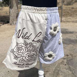 May include: Color-blocked shorts with an elastic waistband. The left side is cream with a brown graphic of roses and the text "Wild Love Roses & Reverie Since 1974". The right side is heather gray and features three dimensional white fabric flowers.