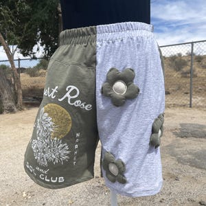 May include: Split-leg shorts featuring an olive green side with a white graphic of cacti, a gold sun, and the text Desert Rose Market and Sol Club, and a light gray side decorated with three dimensional olive green fabric flower appliqués.