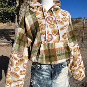 May include: A cropped fleece hoodie with a unique design. The hoodie features a mix of patterns, including green and brown plaid, and a white background with bread illustrations. It has a drawstring hood and a front pocket.